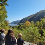 An adult and two children sit on a rocky overlook with a view of Harpers Ferry and the Potomac River below.