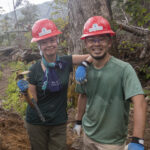 Two volunteers in hardhats stand on a storm-damaged section of the Appalachian Trail with fallen trees.