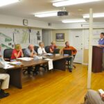 Man stands at podium, presenting to a table of Town Council members.