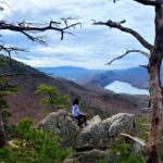 Woman leans on a rock outcropping overlooking a view of mountains and water.